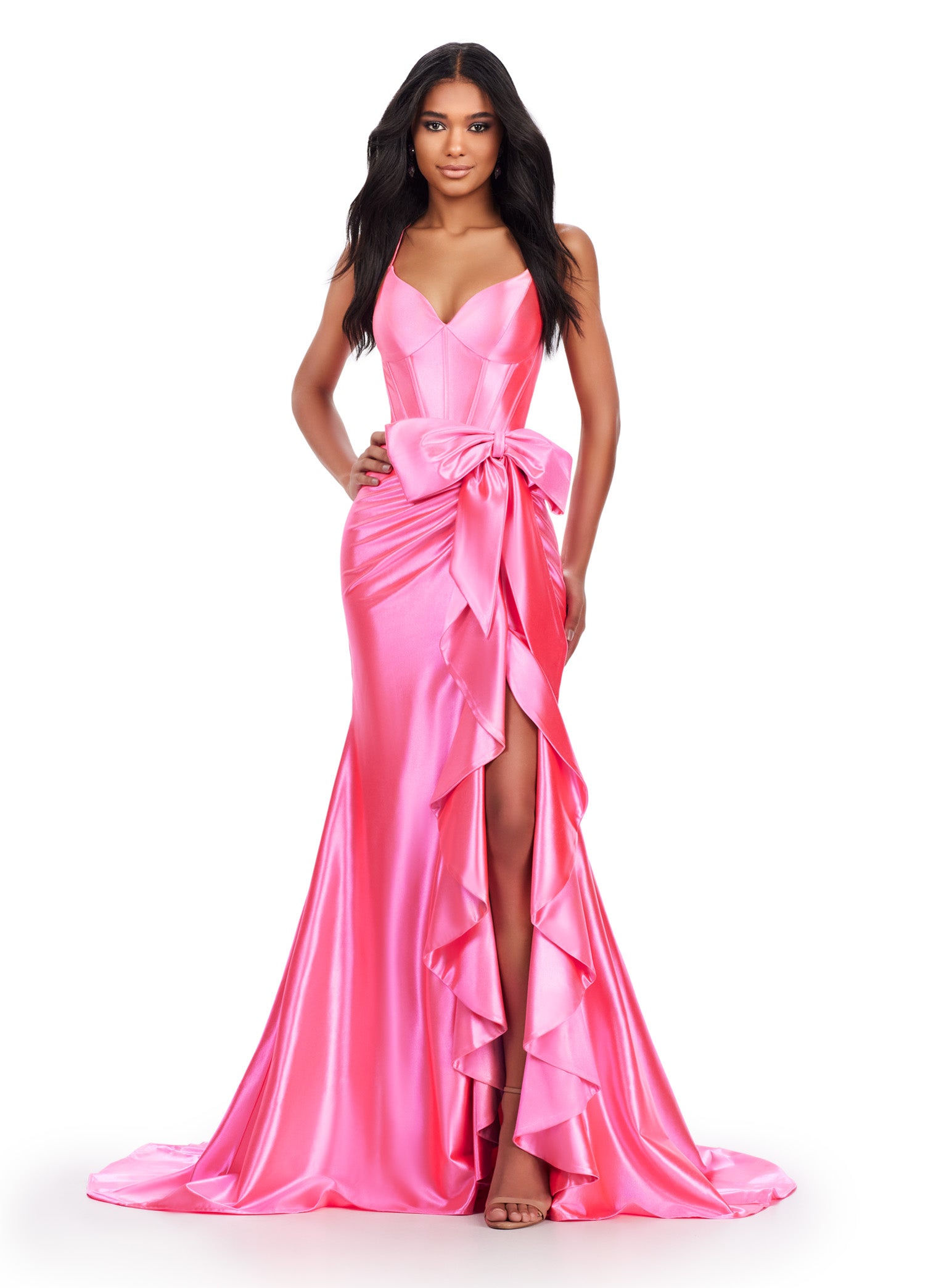 Elegant pink evening gown with a fitted bodice, spaghetti straps, cross back and a large bow accent at the waist. The dress features a cascading ruffle detail along the high slit and flows into a floor-length satin skirt.