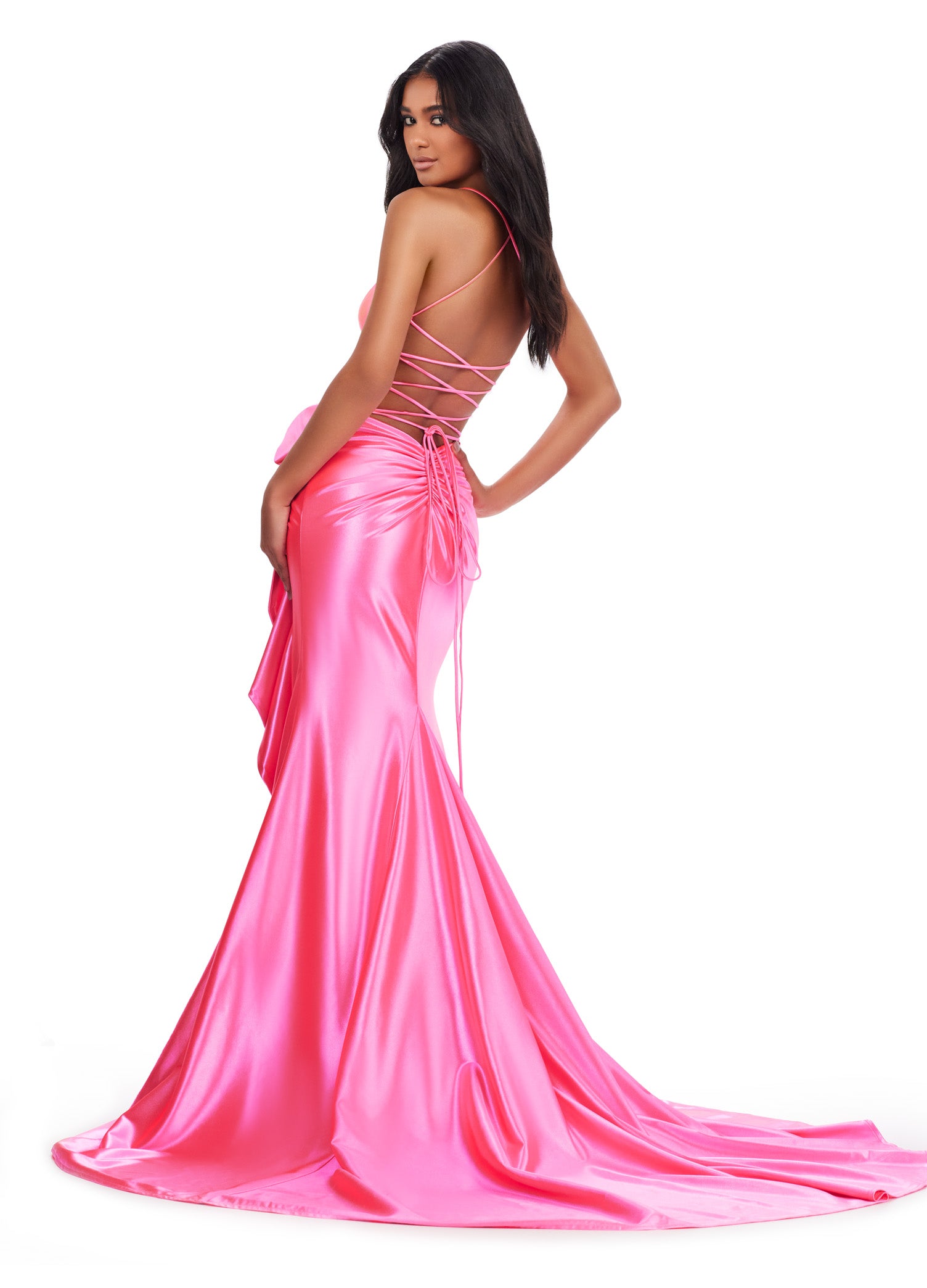 Elegant pink evening gown with a fitted bodice, spaghetti straps, cross back and a large bow accent at the waist. The dress features a cascading ruffle detail along the high slit and flows into a floor-length satin skirt.