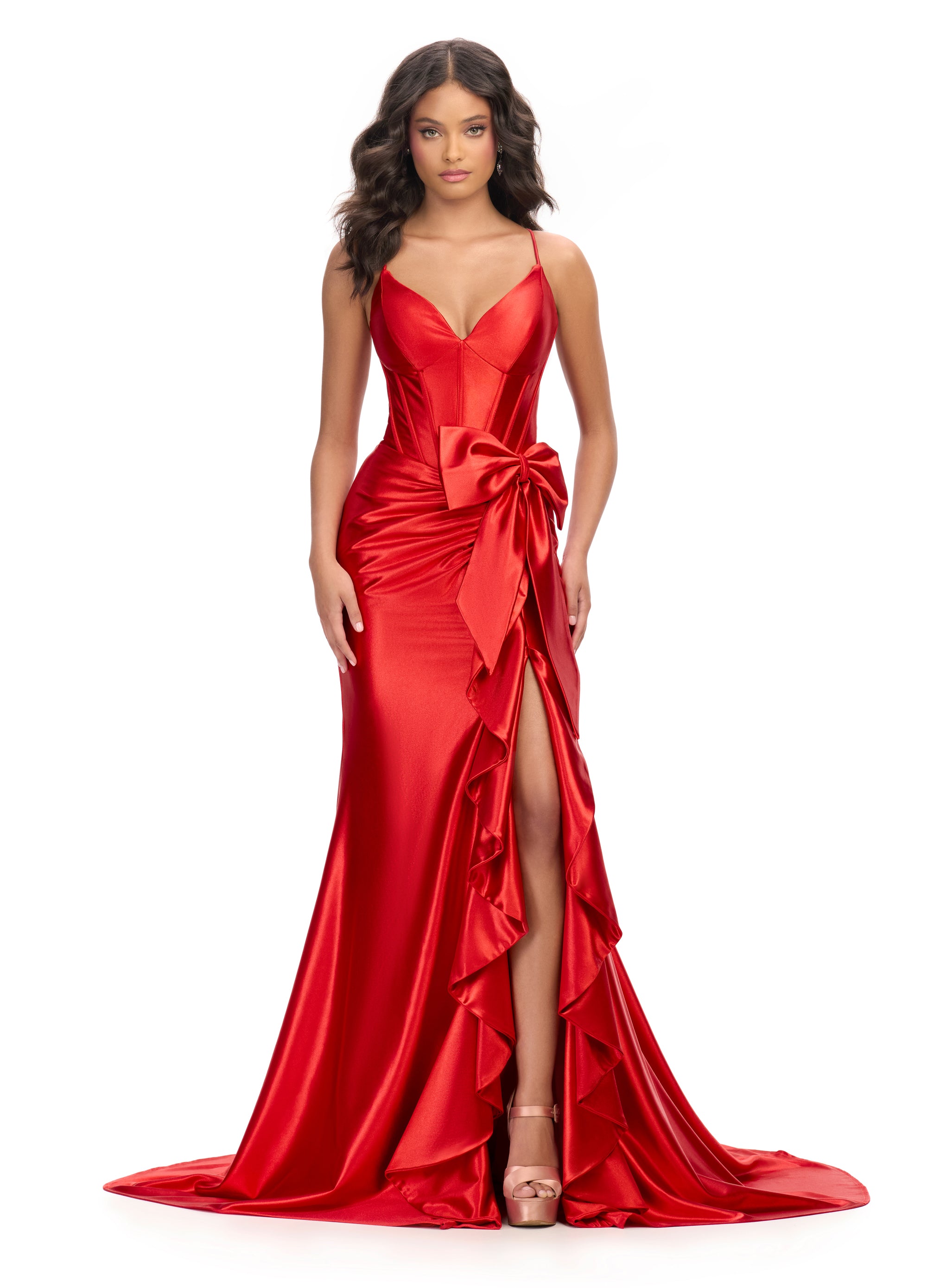 Elegant red evening gown with a fitted bodice, spaghetti straps, cross back and a large bow accent at the waist. The dress features a cascading ruffle detail along the high slit and flows into a floor-length satin skirt.